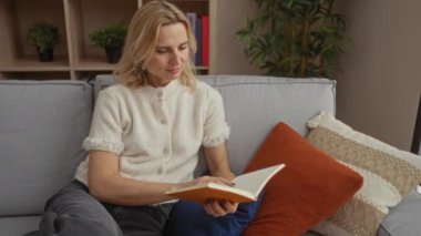 Evdeki rahat bir kanepede kitap okuyan olgun bir kadın. Sıcak bir oturma odasında kitap tutuyor. İçi minderler ve bitkilerle dolu.