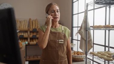 Ekmek ve pasta vitriniyle çevrili önlük giyerken fırında telefonla konuşan bir kadın.