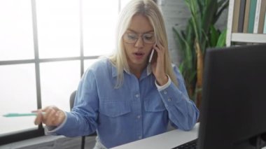 Modern ofiste telefonla konuşan, büyük pencerenin önünde bilgisayarı olan mavi gömlekli ve gözlüklü bir kadın odaklanmış profesyonel bir ortam sergiliyor..