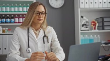 Kadın doktor hastane ofisinde dizüstü bilgisayara bakıyor raflar, şişeler ve arka planda saatle, beyaz önlük ve gözlük takıyor, işyeri gerilimini gösteriyor..