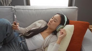 Oturma odasındaki koltukta akıllı telefon ve kulaklıkla dinlenen bir kadın.