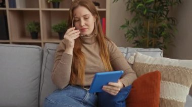 Kadın rahat bir oturma odasında, çiçekli, gözlüklü ve süveterli, rahat ev atmosferi, doğal ışıklandırma, kot pantolon, rahat bir ortam ile rahat bir şekilde otururken tablet okuyor..