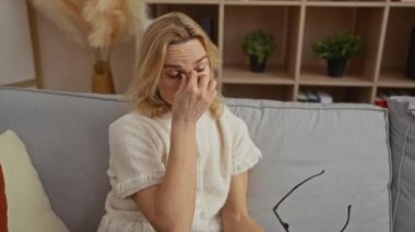 Kapalı bir koltukta oturan yorgun gözleri ovuşturan, gözlük tutan, rahat bir oturma odası ortamında, bej dekorlu ve arka planda bitkiler olan bir kadın sahneyi büyütüyor..