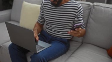 Modern bir oturma odasındaki koltukta oturan bir adam bilgisayarını ve telefonunu kullanıyor.