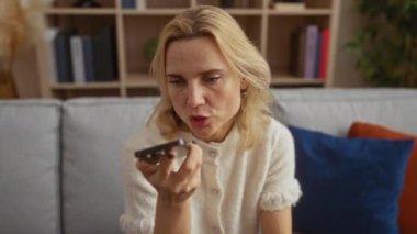 Kapalı mekanda telefonla konuşan bir kadın, rahat modern oturma odasındaki rahat koltukta oturan, rafları ve yastıkları olan, sarı saçlı, rahat ve rahat bir ortam öneren bir kadın..