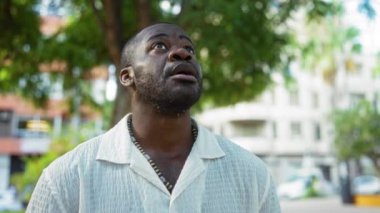 Beyaz gömlekli genç adam arka planda ağaçlar, binalar ve arabalarla şehir parkına bakıyor..