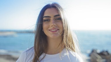 Açık mavi gökyüzüyle deniz kenarında gülümseyen bir kadın canlı bir sahil manzarasında genç güzelliğini ve dingin ifadesini vurguluyor..