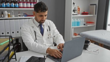 Klinikteki beyaz önlüklü adam laptopta çalışıyor. Etrafı tıbbi ekipman ve dosyalarla çevrili. Profesyonel göreve odaklanmış..