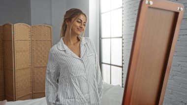 Pijamalarıyla gülümseyen bir kadın, parlak bir yatak odasında sabah ışığını yansıtan büyük bir pencereyle dikilirken..