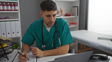 Klinik önlüklü genç adam hastane odasında not alıyor, işine odaklanıyor..