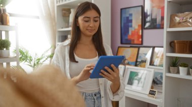Güzel dekore edilmiş bir ev dekorasyonu dükkanında tablet kullanan genç bir kadın. İçinde çeşitli sanat eserleri ve ev bitkileri var.