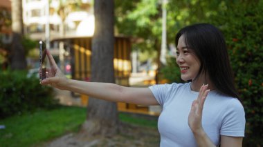 Genç Çinli bir kadın şehir parkında selfie çekiyor, gülümsüyor ve canlı bir şehir ortamında el sallıyor..