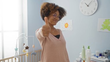 Kıvırcık saçlarıyla gülümseyen bir kadın, bebek beşiğinin yanındaki rahat bir yatak odasında, oyuncaklarıyla ve duvar süslemeleriyle, mutluluk ve sıcaklığını ifade ederek baş parmağını kaldırıyor..