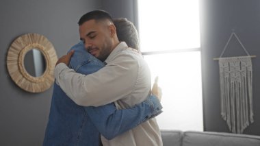 İki yakışıklı adam bir salonda kucaklaşıyor, arkadaşlığı ve bağlantıyı sembolize ediyor, biri kot ceketli, diğeri de bej gömlekli, rahat bir apartman dairesinde..