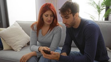 Modern oturma odasındaki kanepede oturan bir çift, akıllı telefondan bir şey gösteren bir kadın, kapalı alanda sıcak bir atmosfer, hem nişanlı, hem de doğal ışık arttırıcı bir ortam.