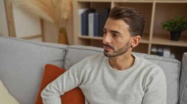 Sakallı genç adam şık oturma odasında koltukta oturuyor, modern iç dekorasyon, kitap rafları ve rahat yastıklarla çevrili iken düşünceli görünüyor..