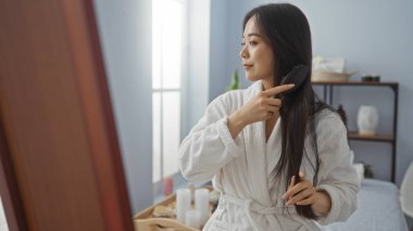 Beyaz bir bornoz giyerken spa ortamında saç tarayan bir kadın, sakin bir ev ortamında rahatlığı, güzelliği ve sağlığını yansıtıyor..