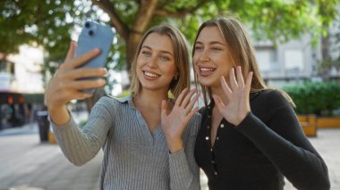 Kardeşler birlikte bir video görüşmesi yapıyor, gülümsüyor ve bir şehir parkında el sallıyor, aile sevgisini ve bağlantısını canlı bir şehir sokak sahnesinde sergiliyorlar..