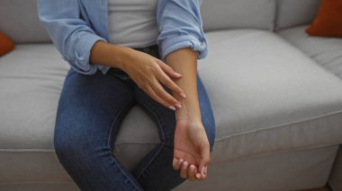 Evde kolunu kaşıyan genç bir kadın, bir salonda oturuyor, günlük mavi gömlek ve kot pantolon giyiyor, rahatsızlık veya kaşıntı tasvir ediyor..