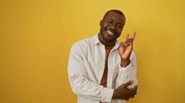 Çekici Afrikalı Amerikalı genç adam gülümsüyor ve izole edilmiş sarı arka plan duvarının üzerinde barış işareti yapıyor.