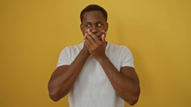 Elleri ağızda olan genç Afrikalı bir adam izole edilmiş sarı arka plana karşı duruyor, sürpriz ya da şok belirtileri gösteriyor, günlük beyaz gömlek giyiyor, güçlü duygular sergiliyor..