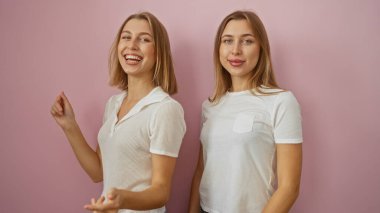 İki gülümseyen sarışın kız kardeş pembe izole bir duvarın önünde dururlar, mutluluk ve aile sevgisini yayarlar, her ikisi de günlük beyaz kıyafetler giyerler, birbirlerine sarılırlar..