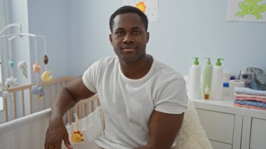 Genç Afrikalı bir adam, beşiği olan rahat bir yatak odasında oturuyor. Etrafı yumuşak dekorlar ve hafif aydınlatmalarla çevrili. Evinde sıcak ve davetkar bir ortam yaratıyor..