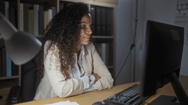 Ofisteki bir masada oturan kıvırcık saçlı kadın, kolları çapraz bilgisayar ekranına bakıyor, beyaz bir ceket giyiyor, arka planda bir sürü dosya var..
