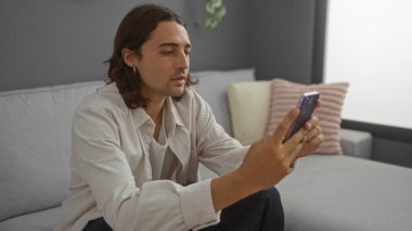 Uzun saçlı, Latin kökenli genç bir adam oturma odasında oturmuş modern bir dairede telefonuna bakıyor..