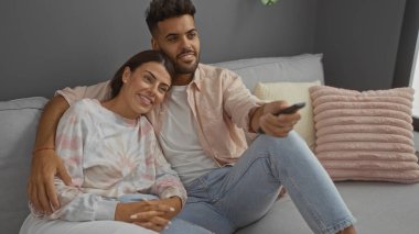 Çift, rahat bir oturma odasında kanepede birlikte rahatlıyor, gülümsüyor ve televizyon seyrediyor, kadın ve erkek yakından oturuyor, evde sıcaklık ve rahatlık hissi yaratıyor.