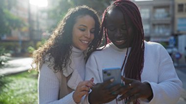Akıllı telefona gülümseyen iki kadın, güneşli bir şehir parkında, canlı bir şehirde arkadaşlığı ve eğlenceyi somutlaştırıyor..