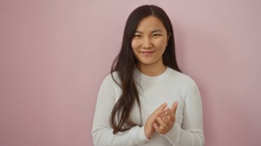 İzole edilmiş pembe arka planda el çırpan bir kadın kendine güvenen ve arkadaş canlısı bir ifadeyle beyaz uzun kollu gömlek giyen bir güzellik sergiliyor.