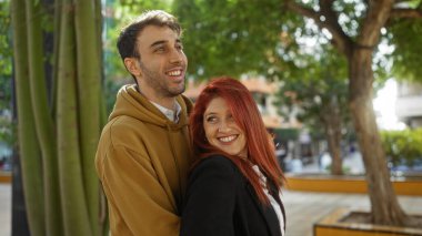 Bir park ortamında gülümseyen adam ve kadın, ağaçlarla çevrili, sevgi ve mutluluk saçan, kentsel bir arka planda görünen.