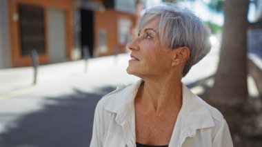 Güneşli bir şehir sokağında kısa saçlı, güzel gri saçlı bir kadın şehir ortamında olgun ve kendinden emin bir tavır sergiliyor..