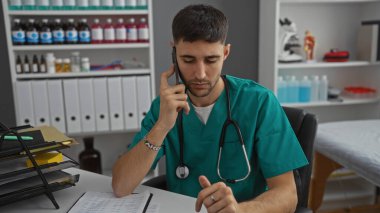 Klinikte önlük giyen genç adam telefonda konuşuyor. Etrafı tıbbi dosyalar ve malzemelerle çevrili. Profesyonel odaklı sağlık ortamını gösteriyor..