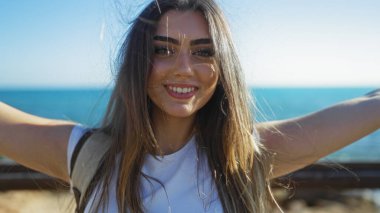 Güneşli bir günde deniz kenarında gülümseyen bir kadın kumsal gezintisinde kendine güvenen esmer bir güzellik sergiliyor..