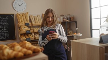 Genç bir kadın fırında akıllı telefon kullanıyor. Etrafı ekmek ve hamur işleriyle çevrili. Arka planda modern bir ev ortamı var.