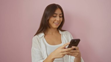 Genç, çekici, İspanyol, akıllı telefonu pembe arka planda kullanırken gülümseyen, gelişigüzel giyinen bir kadın..