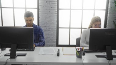 Modern ofisteki bilgisayarlarda çalışan erkek ve kadın, her ikisi de masa başında belgeleri ve ofis malzemelerini olan görevlere odaklanmış, profesyonel bir iş ortamını ön plana çıkarıyorlar..