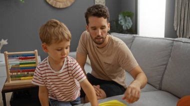 Baba ve oğul evdeki rahat bir oturma odasındaki kanepede birlikte oynuyorlar, çocukluğu ve aile sevgisini yansıtan ahşap bir çerçeveyle renkli bir abaküse odaklanıyorlar..