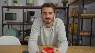 Modern işyeri mutfağında sakallı, kırmızı kupa tutan, arka planda rafları olan, ahşap bir masada yakalanmış genç İspanyol bir adam..