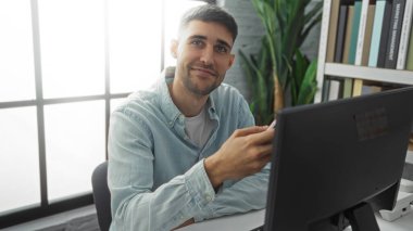 Modern ofiste çalışan genç İspanyol bir adam, bilgisayar kullanıyor, gündelik gömlekler giyiyor, kitaplık ile güvenle gülümsüyor ve arka planda geniş bir pencere var, parlak ve davetkar bir ortam.