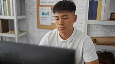 Evinden çalışan genç bir adam modern bir yatak odasında masa başında oturuyor. Duvarında beyaz raflar ve çizelgeler var..