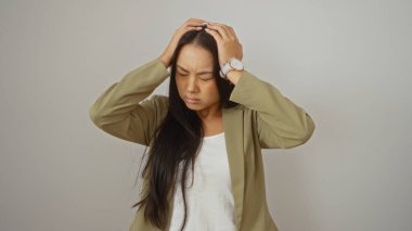 Stresli, uzun saçlı, spor ceket giyen genç Çinli kadın, elleri başının üzerinde, izole edilmiş beyaz bir arka plan üzerinde duruyor.