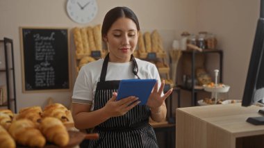 Fırında ekmek ve hamur işleriyle çevrili tablet kullanan genç bir kadın.