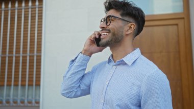 Sakallı genç adam şehir merkezinde telefonda konuşuyor, gözlüklü ve mavi gömlekli, kendine güvenen ve rahat bir hava yayıyor..