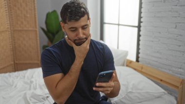 Yatak odasındaki genç adam, modern bir evde elinde akıllı bir telefonla yatağın üzerinde dikkatlice oturur. Pencereden içeri ışık saçar..