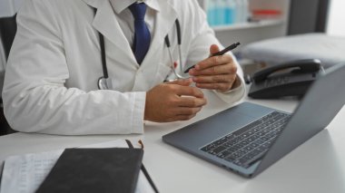 Hastanedeki beyaz önlüklü doktor elinde kalem tutarken ve hasta kayıtlarını tartışırken bilgisayarla çalışıyor..