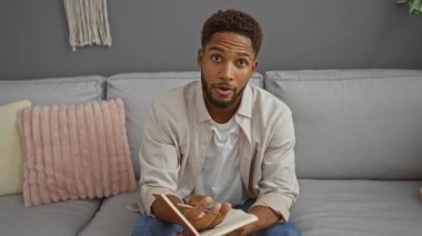 Genç bir adam modern bir oturma odasındaki bir kanepede oturmuş, dekoratif yastıklarla evin içini sergiliyor..