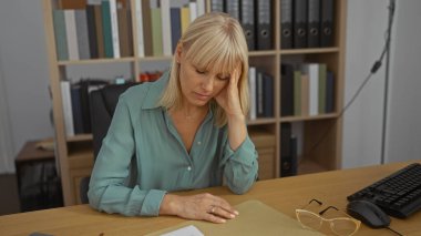 Ofis masasında oturan kadın, iş yerinde eli alnında, evde otururken stresli görünüyor.